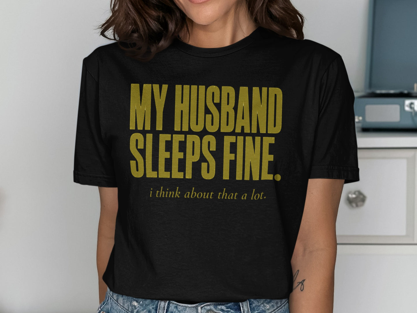 Black t-shirt with gold text reading "MY HUSBAND SLEEPS FINE. i think about that a lot."
