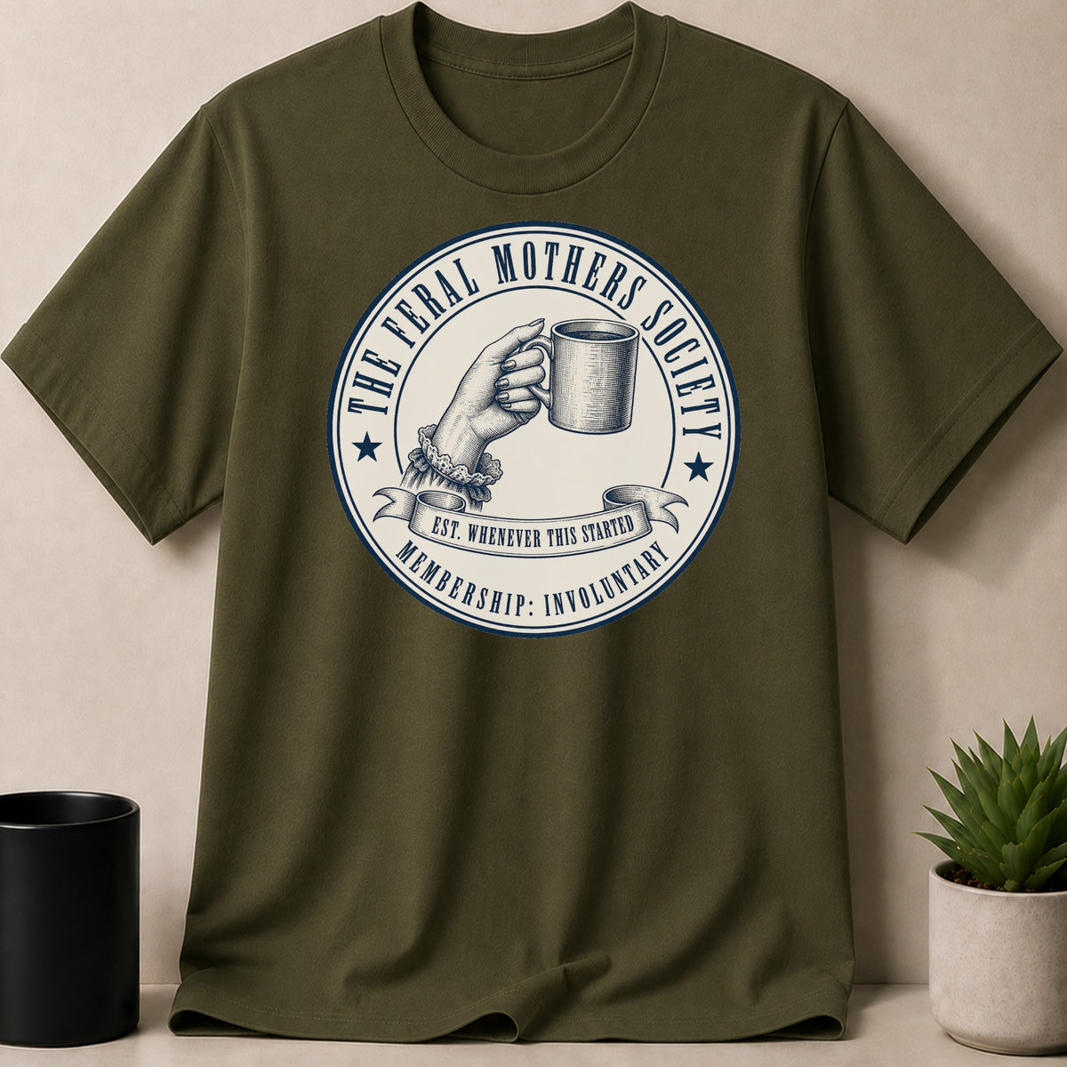 Olive t-shirt with "The Feral Mothers Society" circular emblem featuring a hand holding a mug.