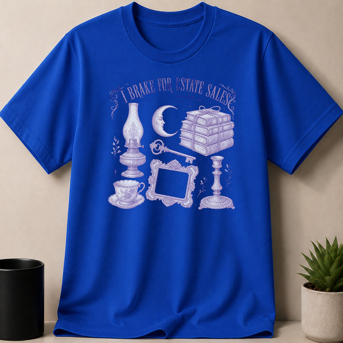 Blue t-shirt with purple line art of lamp, books, moon, frame, key, cup, and candlestick.