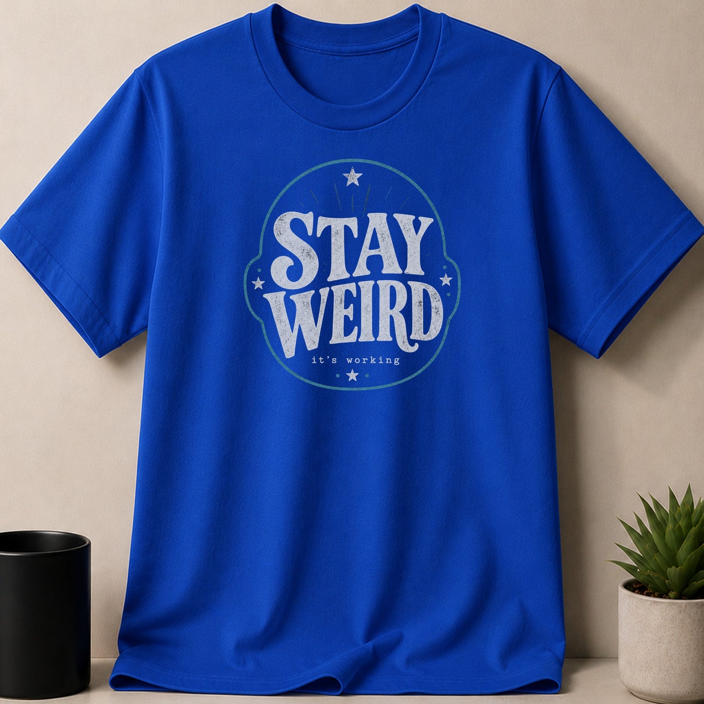 Royal blue t-shirt with "STAY WEIRD it's working" text in distressed white lettering.