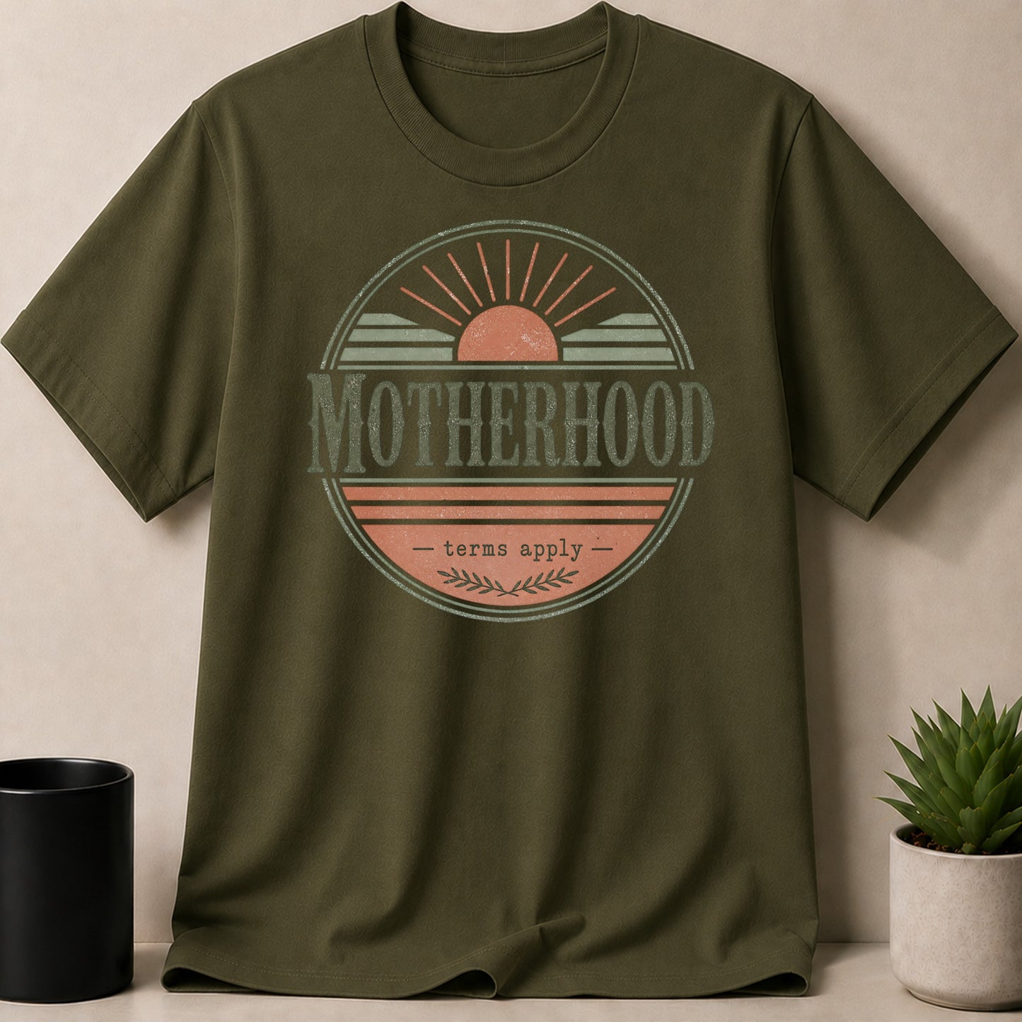 Olive green t-shirt with a circular graphic stating "MOTHERHOOD - terms apply".