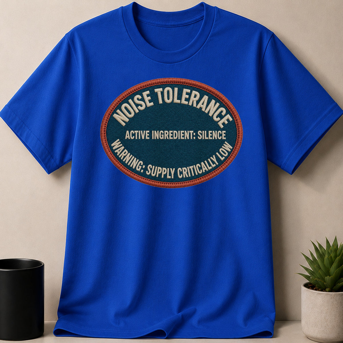 Blue t-shirt with embroidered oval patch stating "Noise Tolerance Active Ingredient: Silence Warning: Supply Critically Low".