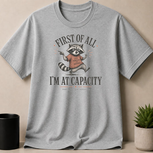 Gray t-shirt with raccoon graphic that reads "First of all I'm at capacity."