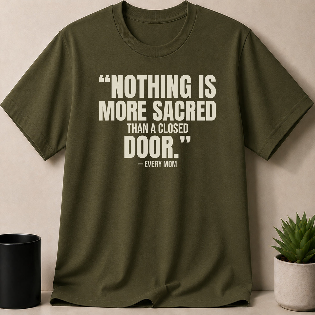 An olive green t-shirt with white text reading "NOTHING IS MORE SACRED THAN A CLOSED DOOR. – EVERY MOM" displayed centrally.