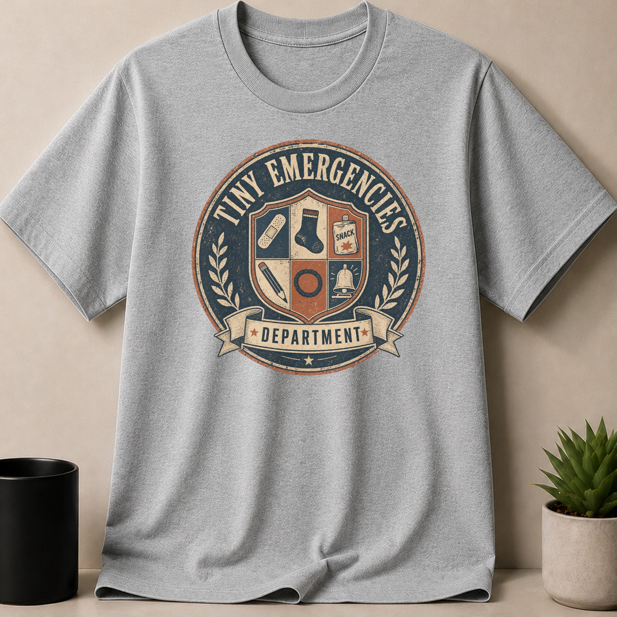 Grey t-shirt with "Tiny Emergencies Department" emblem featuring band-Aid, sock, snack, pencil, and bell.