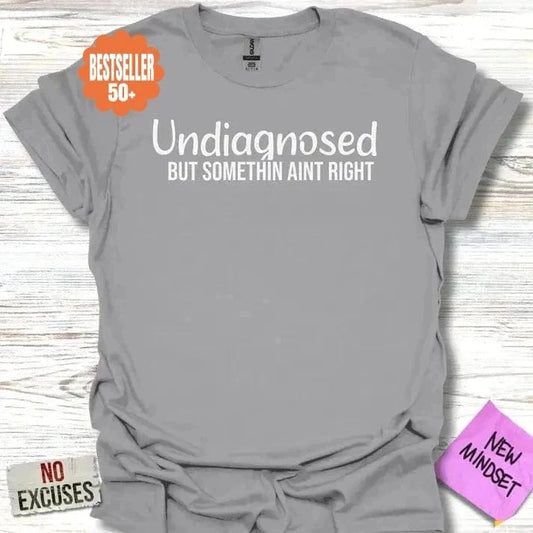 Gray t-shirt with text 'Undiagnosed BUT SOMETHIN AINT RIGHT' on a wooden surface.