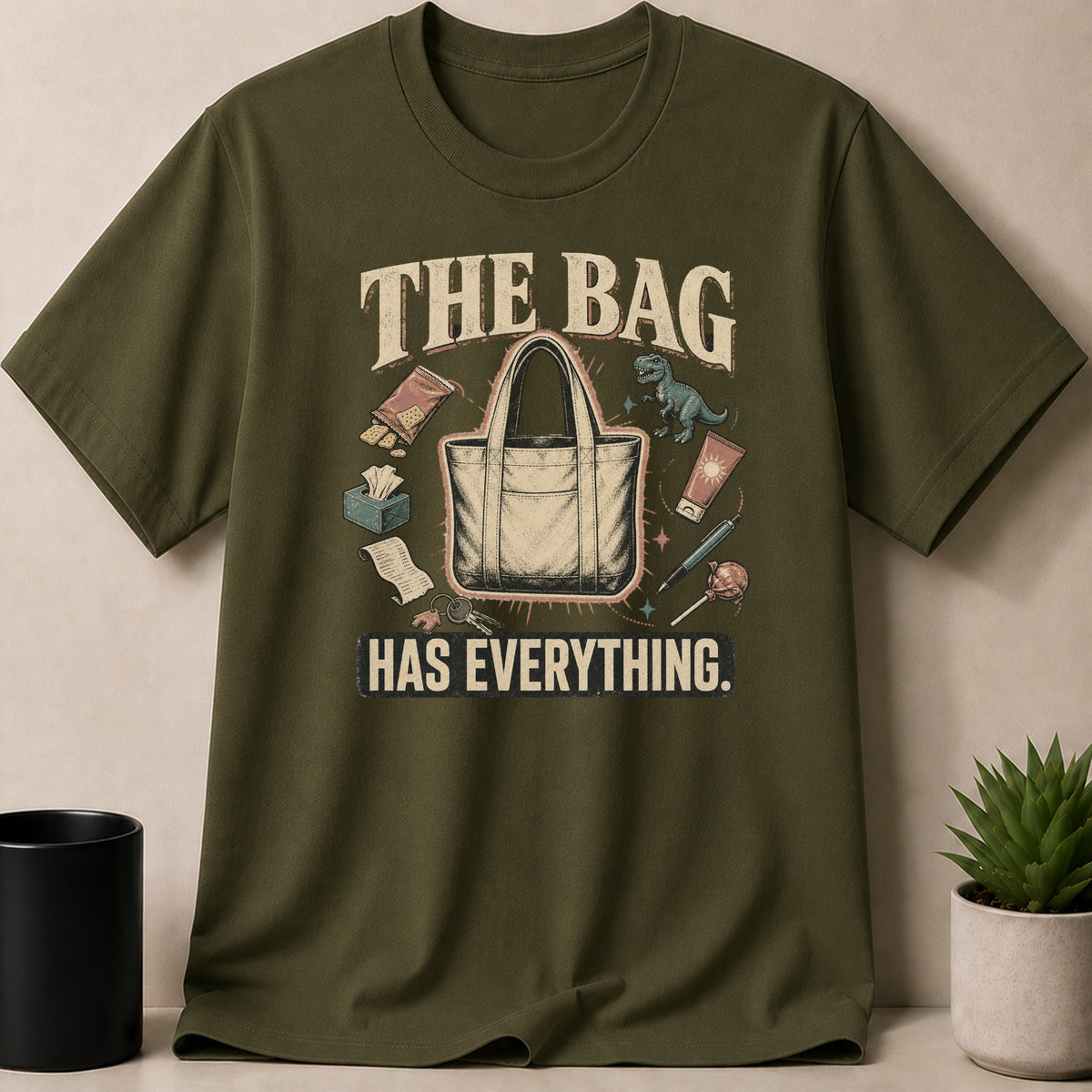 Olive t-shirt with "The bag has everything" graphic featuring a tote bag and various items.