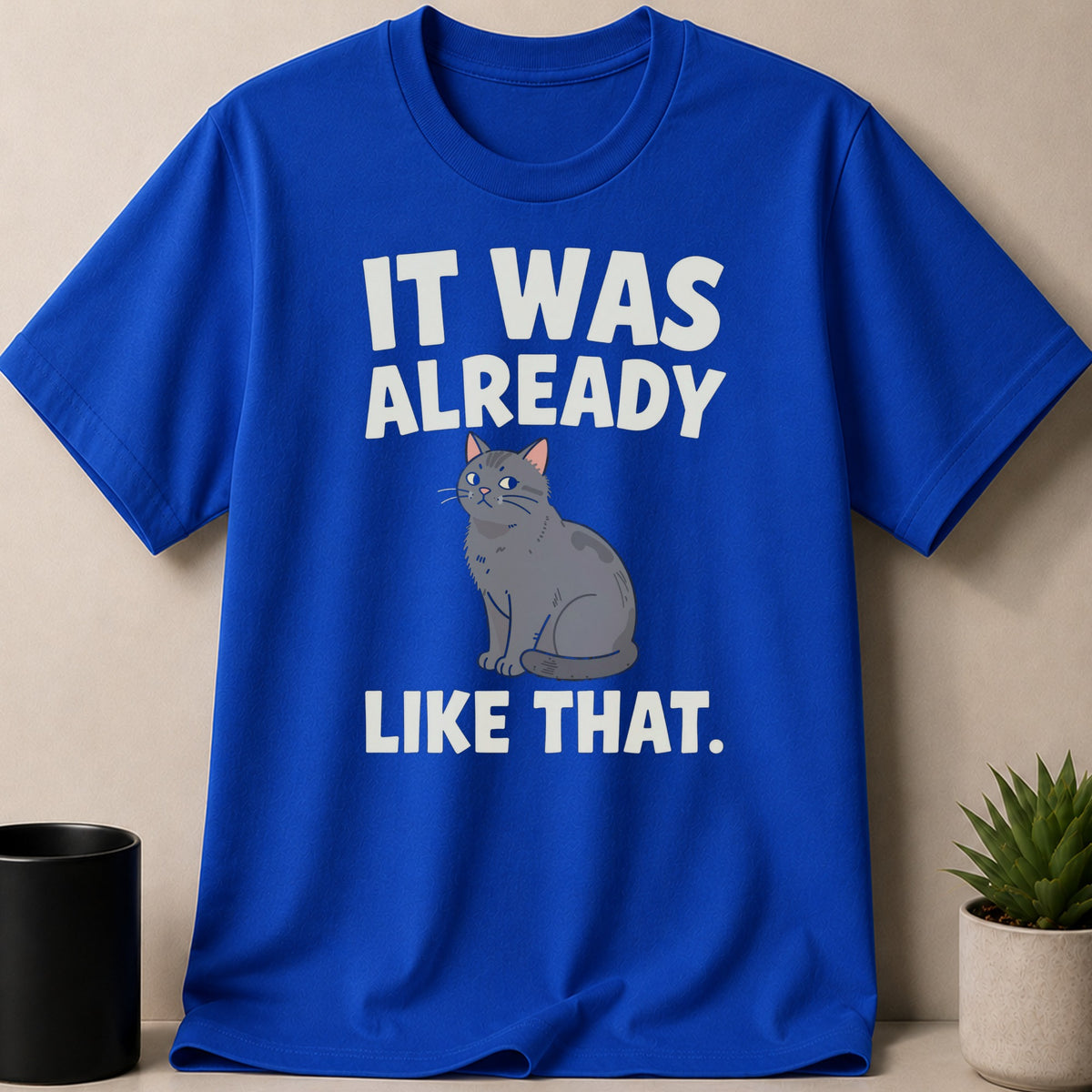 Bright blue crewneck t-shirt featuring white text that reads IT WAS ALREADY LIKE THAT and a grey cat illustration.