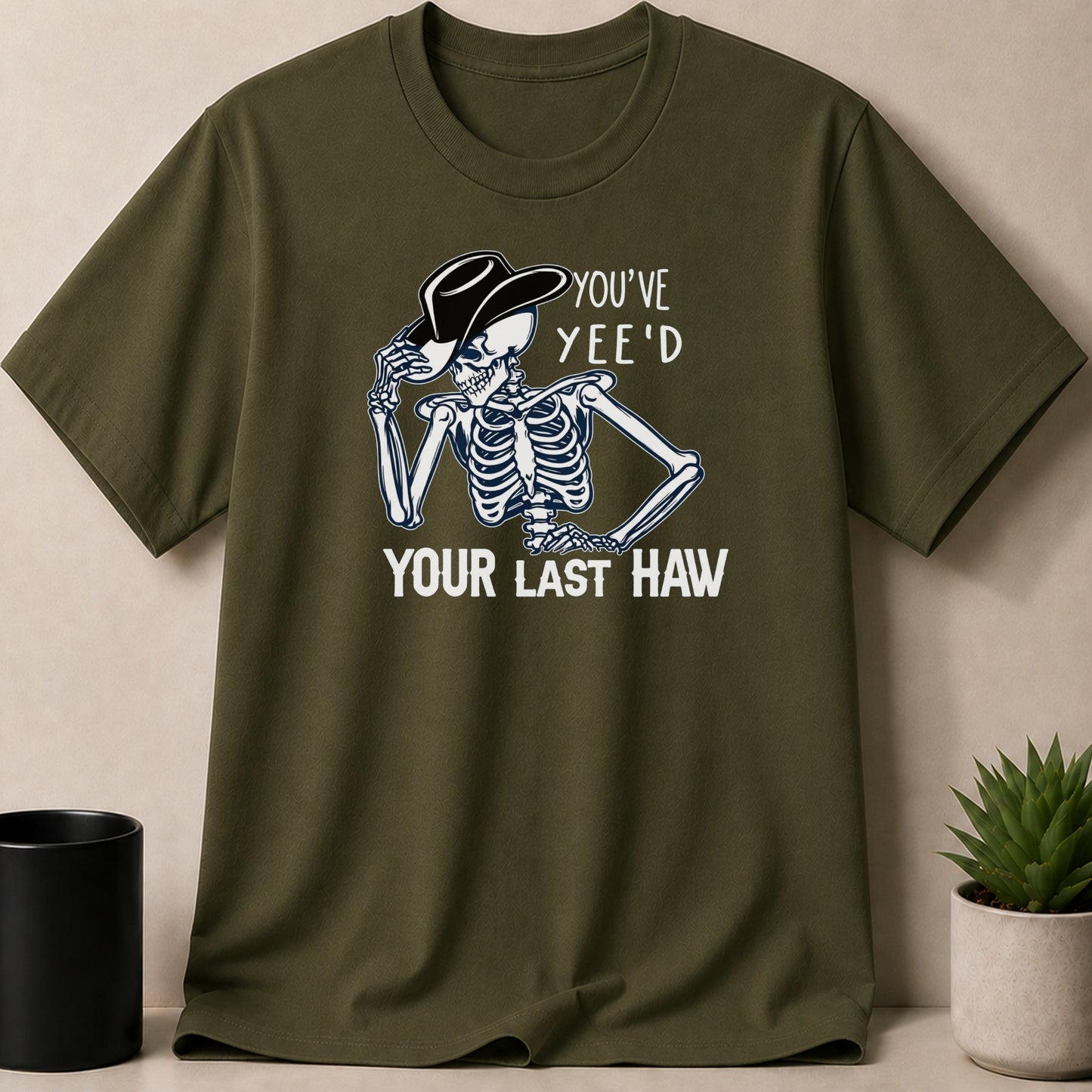 Olive green t-shirt with skeleton wearing cowboy hat and text "YOU'VE YEE'D YOUR LAST HAW".