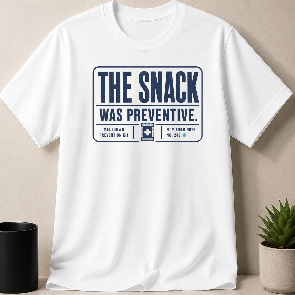 White t-shirt with navy text reading "The snack was preventive. Meltdown prevention kit. Mom field note no. 247."