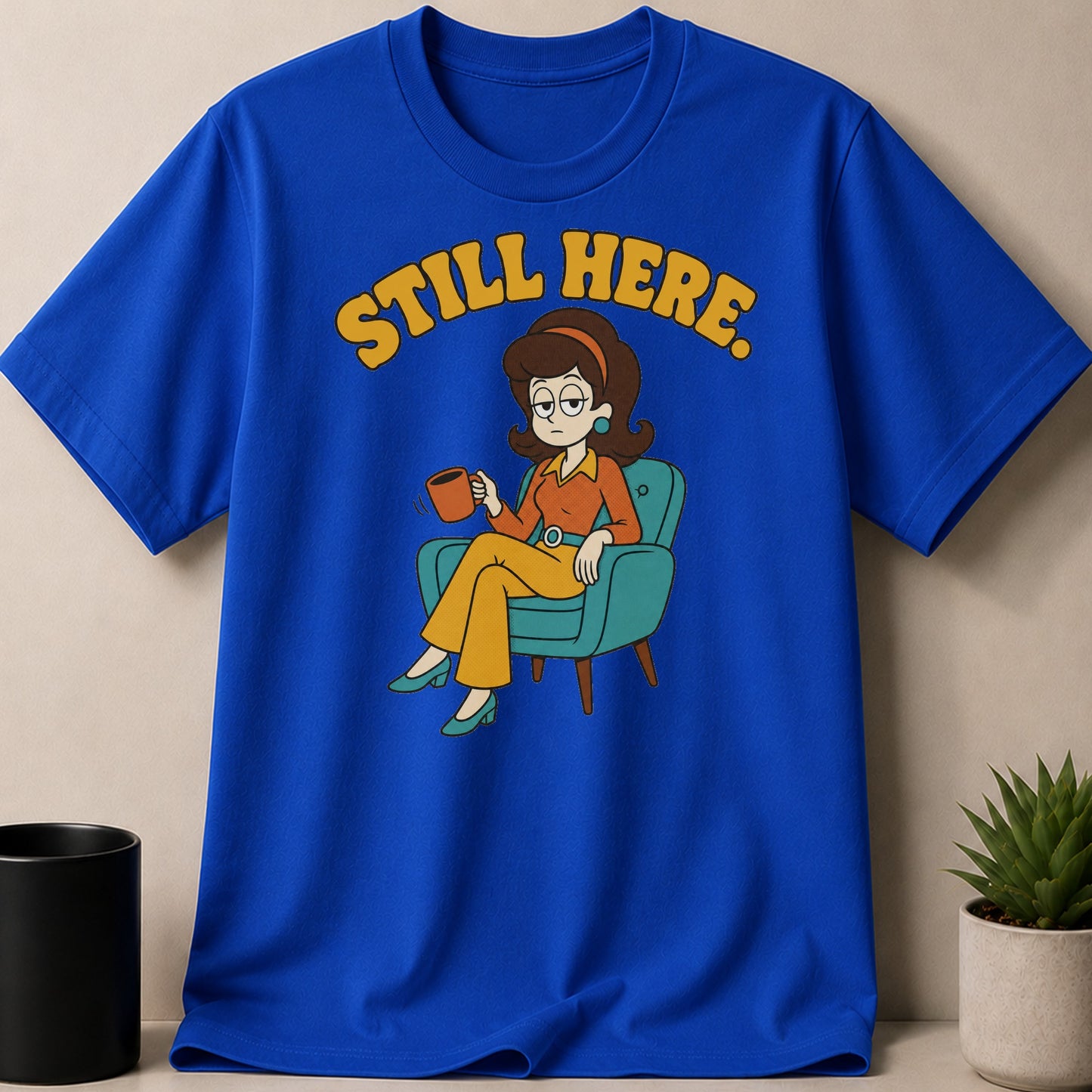 Blue t-shirt with "STILL HERE." text and cartoon woman sitting in chair with coffee.