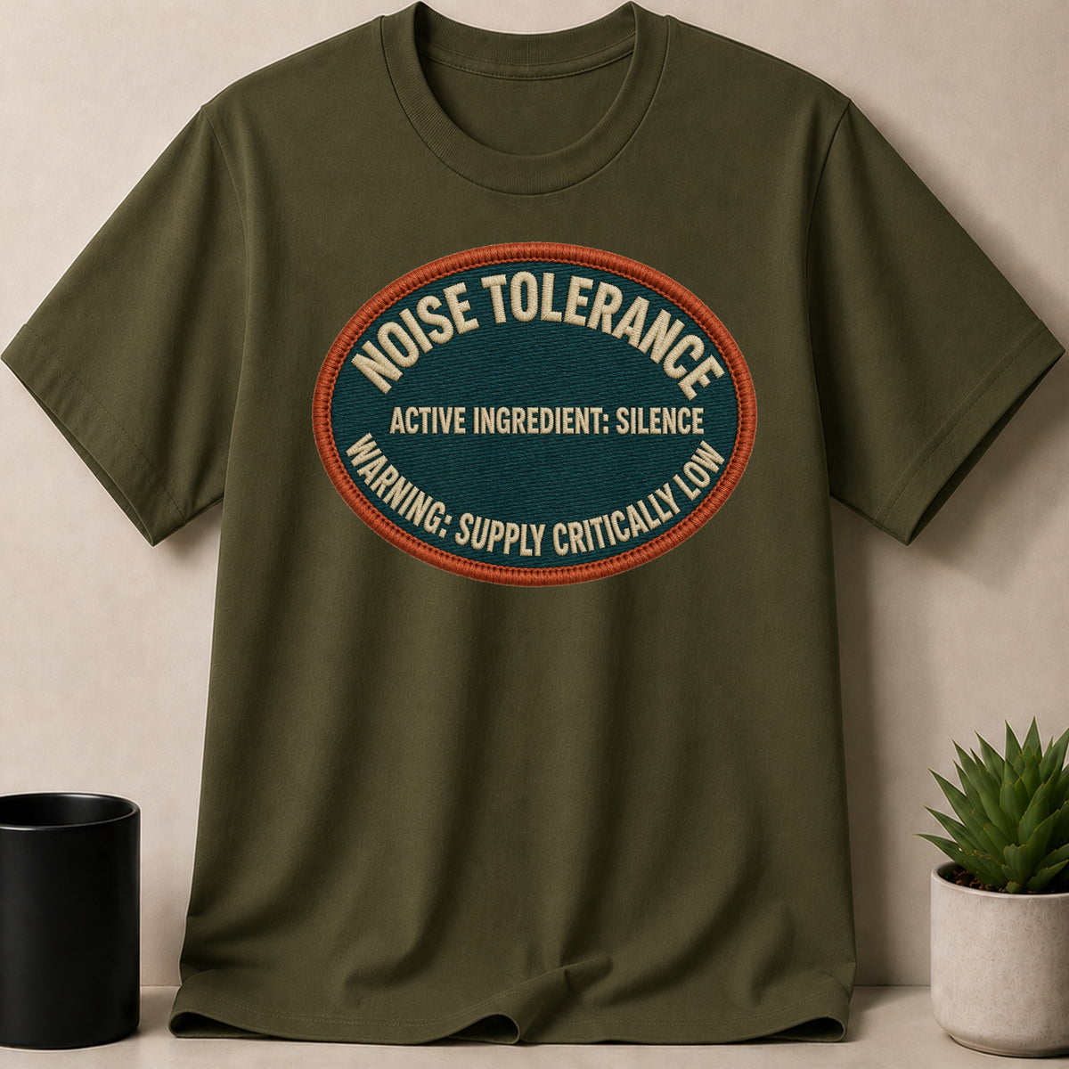 Olive green t-shirt with "Noise Tolerance" patch: "Active Ingredient: Silence. Warning: Supply Critically Low."
