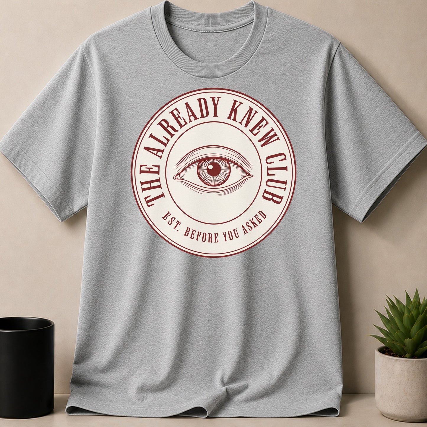 Grey t-shirt with circular emblem featuring an eye and text "The Already Knew Club Est. Before You Asked".