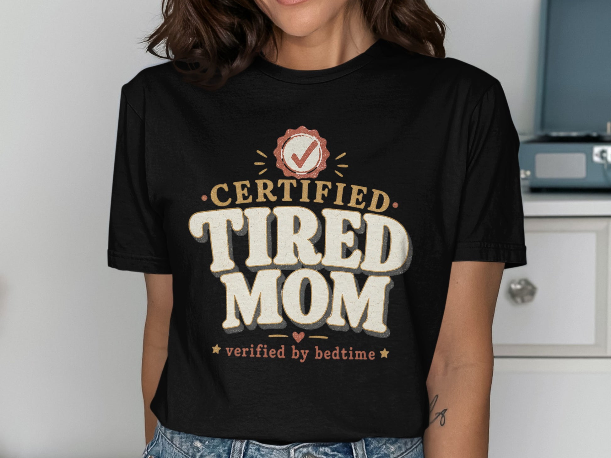 Black t-shirt with "Certified Tired Mom verified by bedtime" text and graphics.