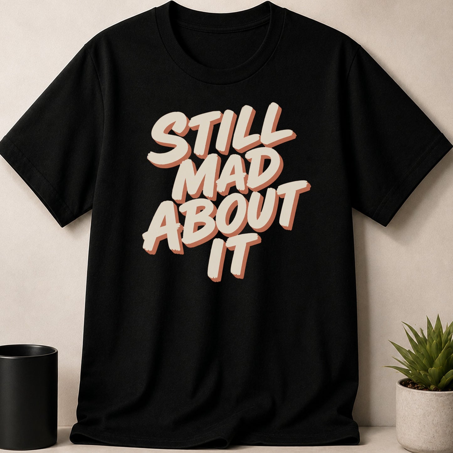 Black t-shirt with "STILL MAD ABOUT IT" text in cream and orange lettering.