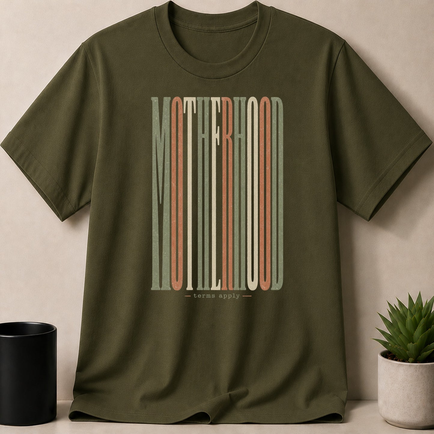 Olive green t-shirt with "MOTHERHOOD - terms apply" in colorful vertical striped lettering.