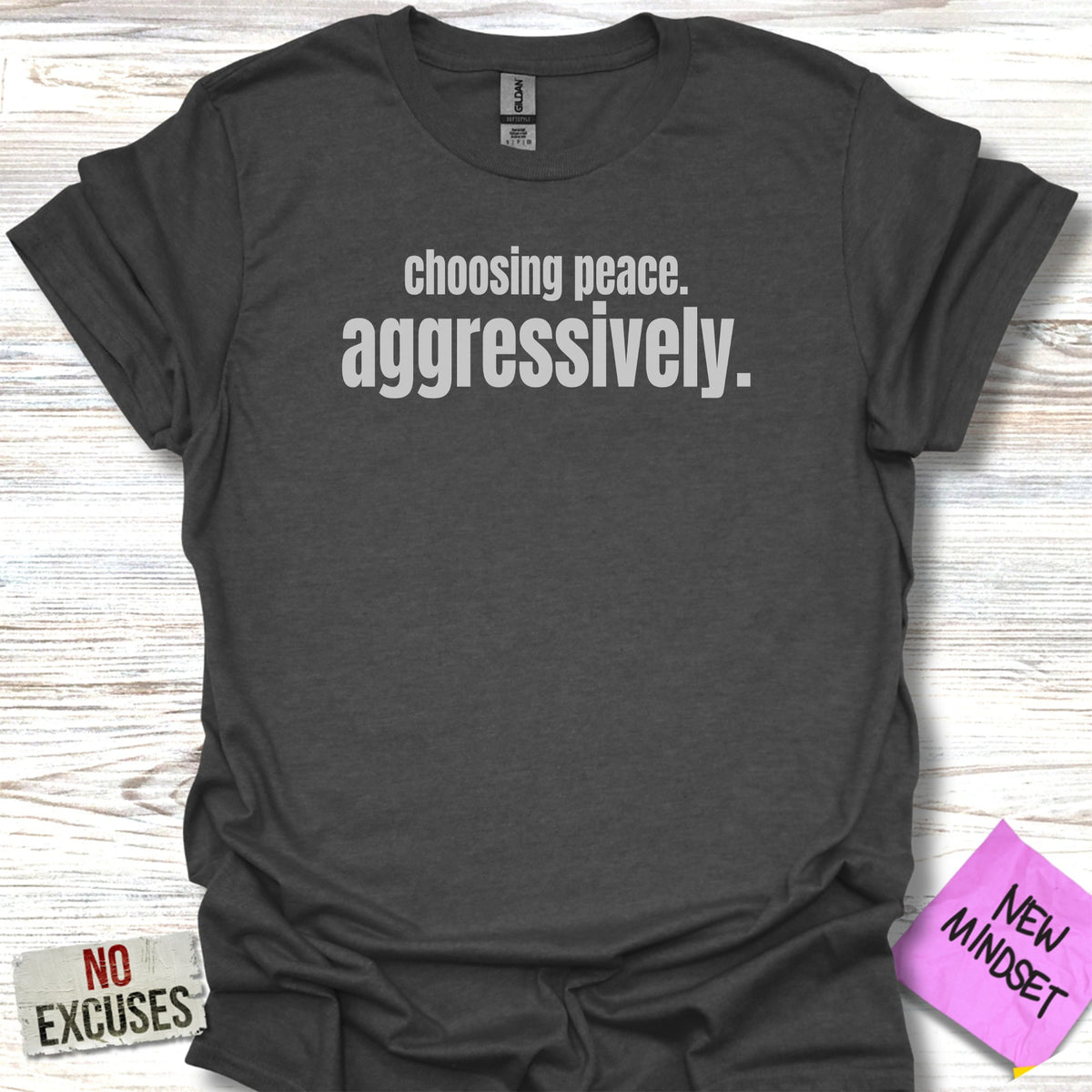 Dark grey t-shirt with white text "choosing peace. aggressively." and "NO EXCUSES" in red.