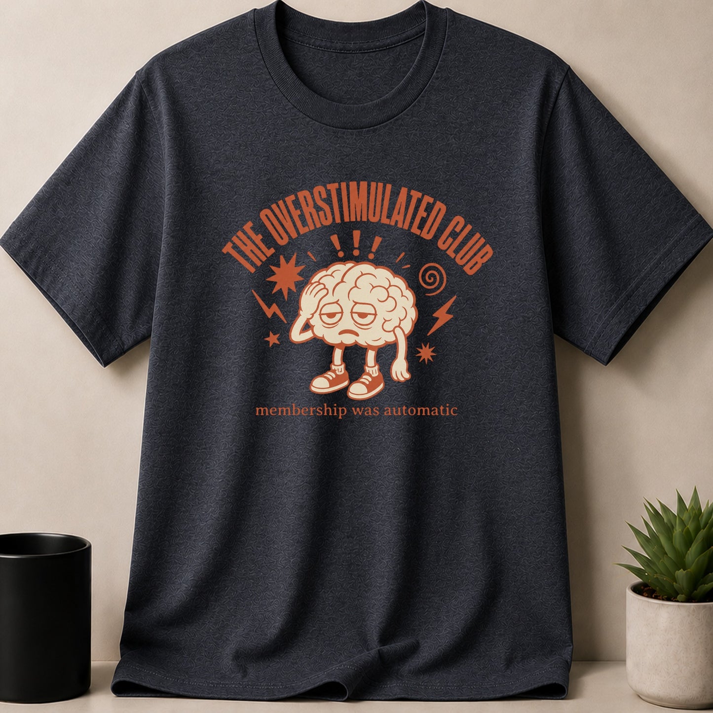 Dark t-shirt with an illustration of a tired brain and text "THE OVERSTIMULATED CLUB" and "membership was automatic".