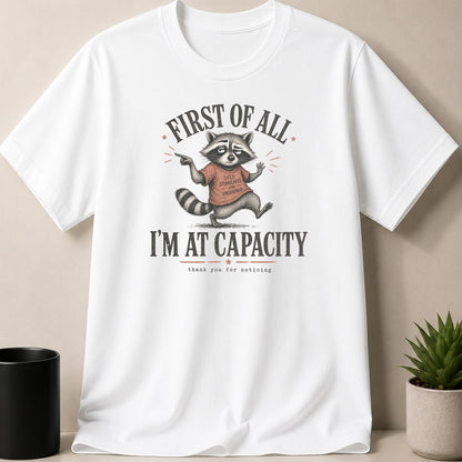 White t-shirt with raccoon illustration saying "First of all, I'm at capacity."
