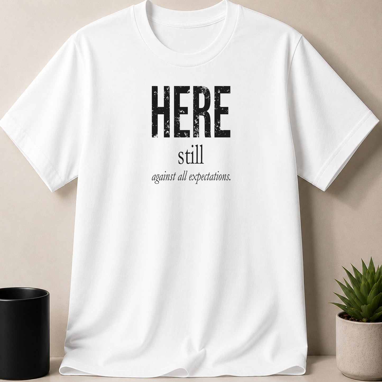 White t-shirt with black text "HERE still against all expectations."