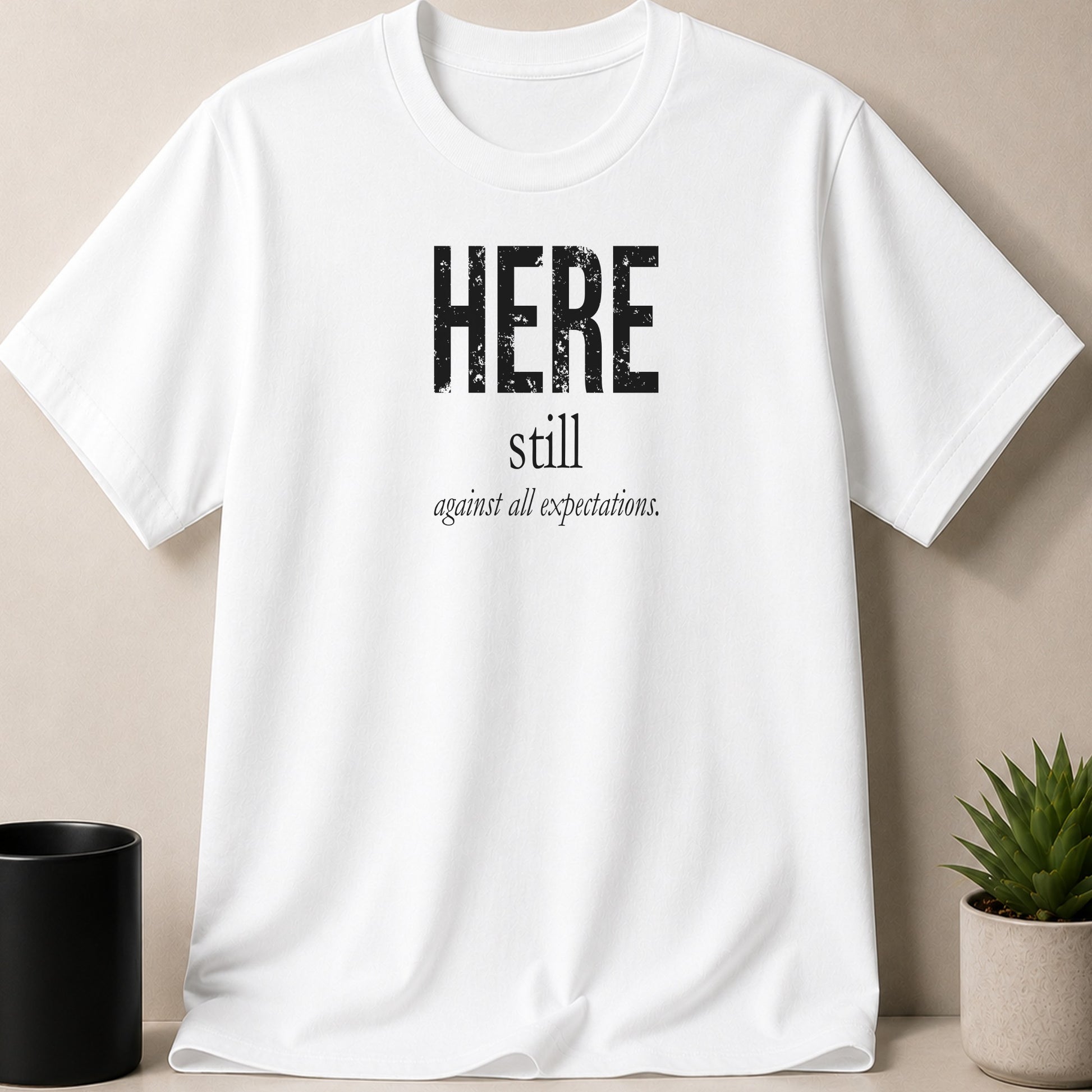 White t-shirt with black text "HERE still against all expectations."