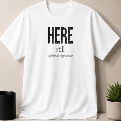 White t-shirt with black text "HERE still against all expectations."