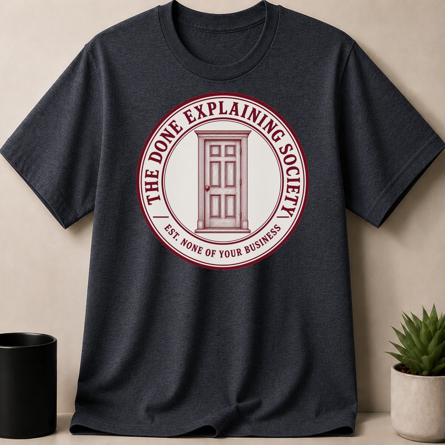 Dark t-shirt with a circular logo featuring a door and the text "THE DONE EXPLAINING SOCIETY EST. NONE OF YOUR BUSINESS".